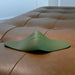 The Gender X Pleasure Pad Rechargeable Grinding Pad & Stroker, featuring a green, curved silicone design with smooth ridges and a power button symbol, rests on a tan tufted leather surface. The device showcases a modern minimalist look.