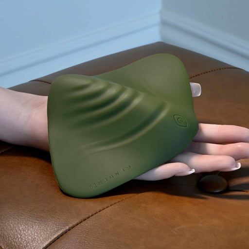 A hand holds the Gender X Pleasure Pad Rechargeable Grinding Pad & Stroker—a green, waterproof silicone pad with ridges and a button—on a brown leather surface.