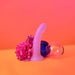 The Glitzy Smooth 7 inch Pink Sparkle Silicone Dildo with Suction Cup stands upright beside pink flowers and a blue glass on a vibrant pink and orange background.