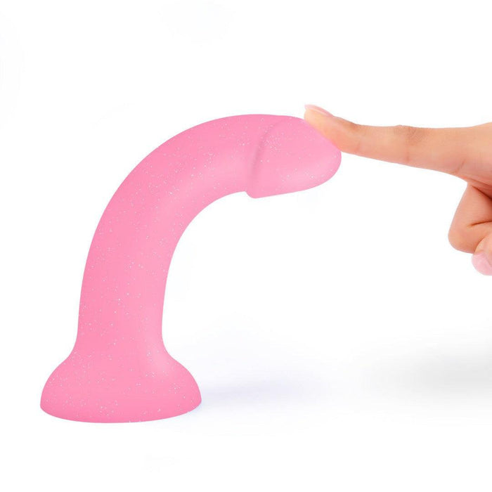 A hand gently presses the tip of the Glitzy Smooth 7 inch Pink Sparkle Silicone Dildo with Suction Cup, featuring a curved design and flat base, set against a plain white background.