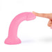 A hand gently presses the tip of the Glitzy Smooth 7 inch Pink Sparkle Silicone Dildo with Suction Cup, featuring a curved design and flat base, set against a plain white background.