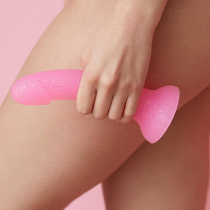A person holds the Glitzy Smooth 7 inch Pink Sparkle Silicone Dildo with Suction Cup near their upper thigh against a matching pink background.