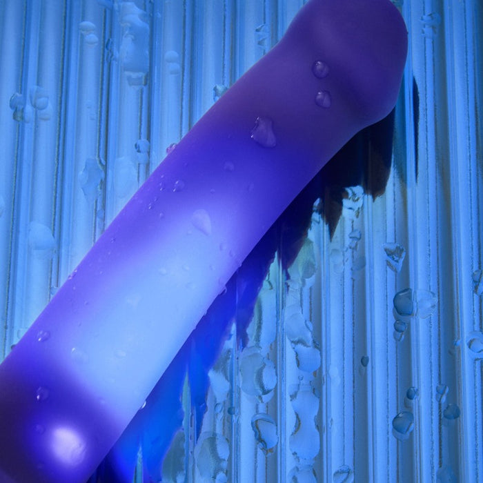 The Glow Light Up Blue LED 7 Inch Silicone Dildo, featuring a glowing blue silicone surface with water droplets, rests on a wet, ridged blue background; it’s strap-on harness compatible.