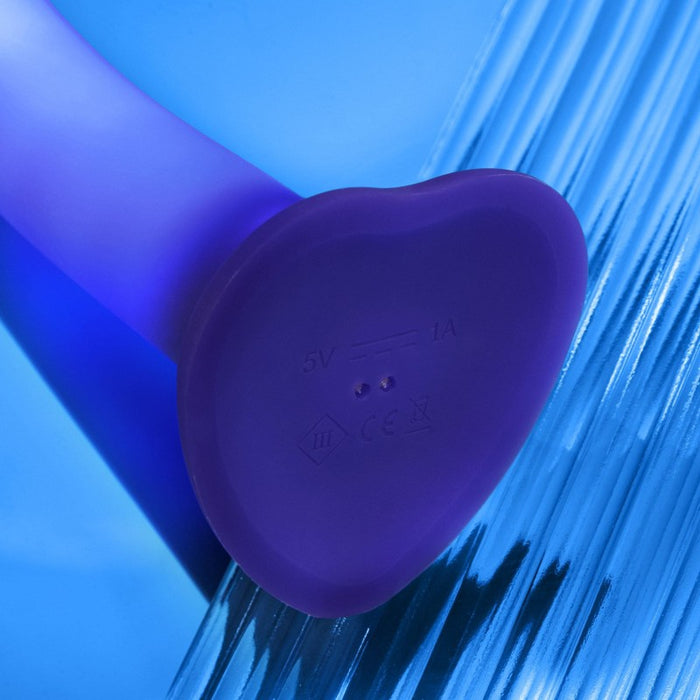 Close-up of the Glow Light Up Blue LED 7 Inch Silicone Dildo, featuring a heart-shaped base, small charging indicators, and LED glow. Its purple silicone design is strap-on harness compatible, set against a blue, glass-textured background.