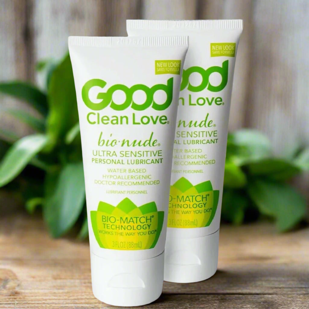 Two tubes of Good Clean Love BioNude Ultra Sensitive Personal Lubricant stand upright on a wooden surface with green leaves blurred in the background. The tubes are white with green and yellow text and accents.