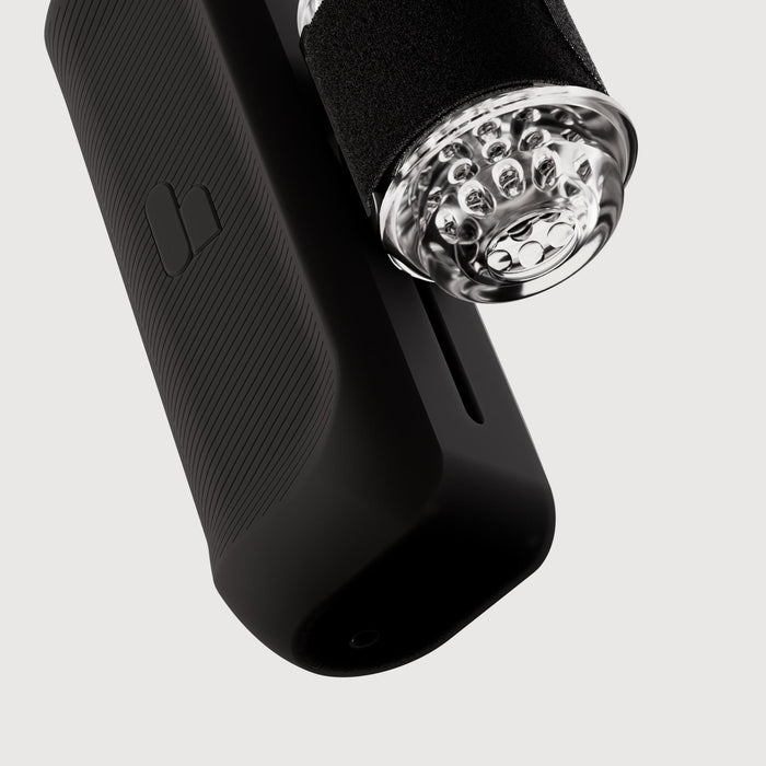 A close-up of The Handy 2 Interactive Automatic Masturbator, a black textured device with a transparent cylindrical attachment featuring multiple small round parts inside, shown against a plain white background.