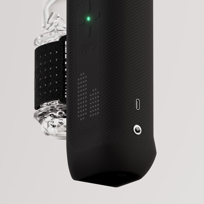 A close-up of The Handy 2 Interactive Automatic Masturbator, a sleek black male sex toy featuring a glowing green indicator, tactile buttons, patterned grip, and an attached transparent container secured by a black strap on a light background.