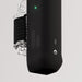 A close-up of The Handy 2 Interactive Automatic Masturbator, a sleek black male sex toy featuring a glowing green indicator, tactile buttons, patterned grip, and an attached transparent container secured by a black strap on a light background.