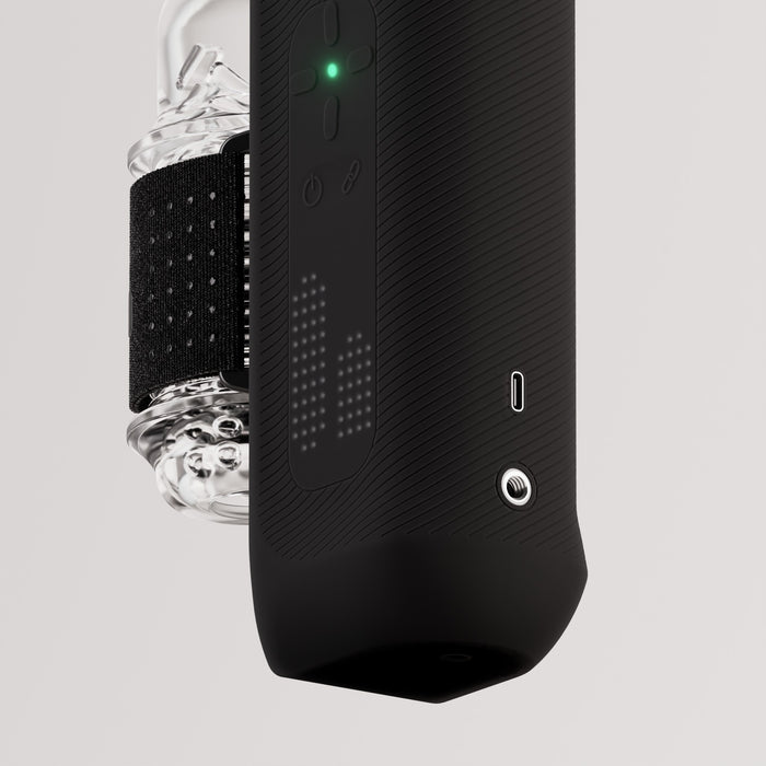 Close-up of a black, cylindrical portable speaker with display, USB-C port, and 3.5mm jack; The Handy 2 Interactive Automatic Masturbator is attached to the side along with a transparent water bottle featuring a black strap.