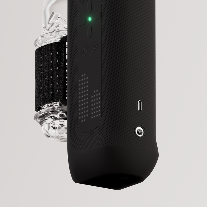 A close-up of The Handy 2 Interactive Automatic Masturbator, a sleek black male sex toy featuring a glowing green indicator, tactile buttons, patterned grip, and an attached transparent container secured by a black strap on a light background.