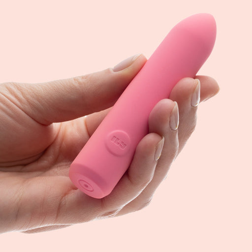 A hand holds the Ily Squishy Classic Silicone Bullet Vibrator—a pink, cylindrical, rechargeable silicone sex toy with a rounded tip—set against a light pink background.
