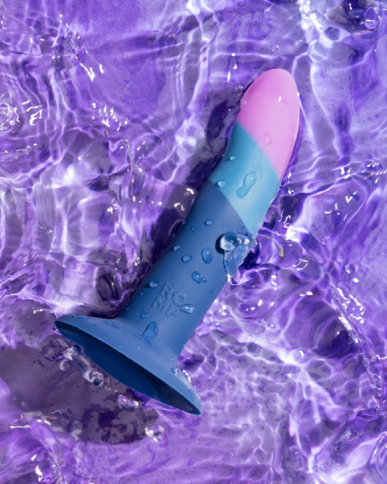 Colorful Lovehoney Romp Piccolo First Time Pegging Kit - Strap on Harness + Dildo partially submerged in water with swirling purple hues around it, reflecting shimmering light.