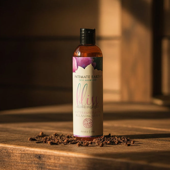 A bottle of Intimate Earth Bliss Anal Relaxer Lubricant with Clove Oil sits on a wooden surface, surrounded by scattered cloves and bathed in warm sunlight.