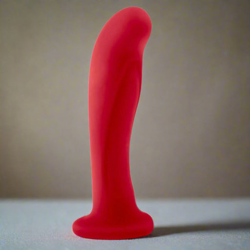 The Jezebel Sleek Curved 6 Inch Red Silicone G-Spot Dildo, featuring a flared suction cup base, stands upright on a light surface against a neutral background.