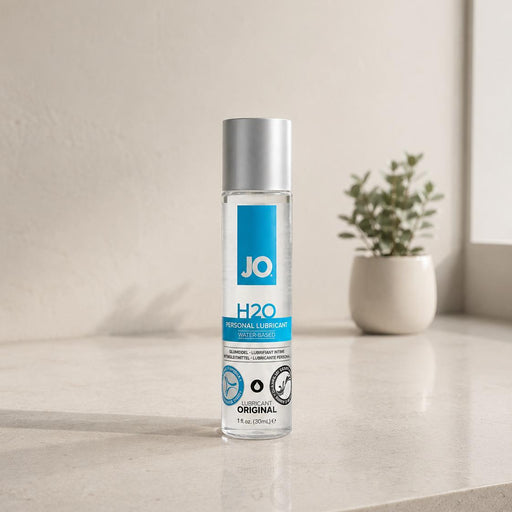 A clear bottle of Jo H2O Original Water Based Lubricant—with a silver cap—sits on a beige countertop. Made with plant-based glycerin and compatible with latex condoms, it’s seen near a small potted plant and softly lit window in the background.