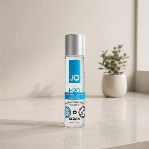 A clear bottle of Jo H2O Original Water Based Lubricant—with a silver cap—sits on a beige countertop. Made with plant-based glycerin and compatible with latex condoms, it’s seen near a small potted plant and softly lit window in the background.