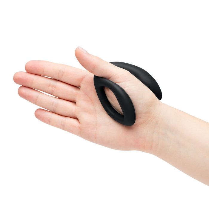 A hand holds a small, black, oval-shaped silicone ring with a curved design from the Womanizer x We-Vibe Lust & Luxury Collection against a white background.