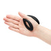 A hand holds a small, black, oval-shaped silicone ring with a curved design from the Womanizer x We-Vibe Lust & Luxury Collection against a white background.