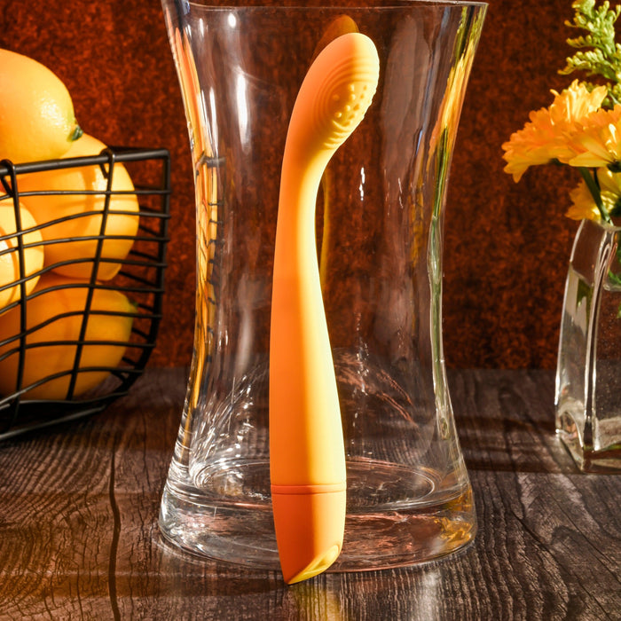 A yellow, ergonomically designed Lemon Squeeze Slim G-Spot Beginner Vibrator by Evolved Novelties stands upright inside a clear glass vase. Made of silky silicone for G-spot stimulation, the scene is set on a wooden surface with a wire basket containing oranges and another glass vase holding yellow flowers in the background.