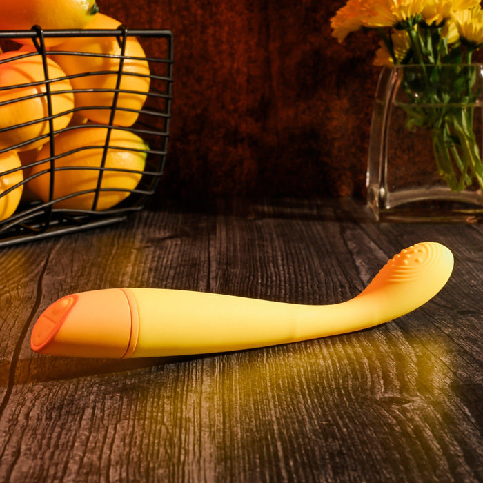 A Lemon Squeeze Slim G-Spot Beginner Vibrator - Yellow by Evolved Novelties, made of silky silicone and waterproof, is displayed on a wooden surface. In the background, there is a wire basket filled with lemons on the left and a glass vase containing yellow flowers on the right. The scene is warmly lit, creating a cozy ambiance perfect for G-spot stimulation.