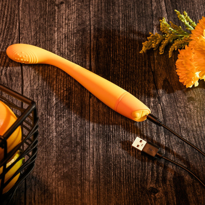 The Lemon Squeeze Slim G-Spot Beginner Vibrator in yellow from Evolved Novelties, crafted from silky silicone, is connected to a USB charger on a rustic wooden surface. Nearby, a black metal basket and yellow flowers are visible. The scene is warmly lit, creating a cozy atmosphere.
