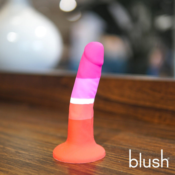 The Lesbian Pride Small 5.5 Inch Silicone Suction Cup Dildo, featuring pink, white, and red stripes and a silky finish, stands upright on a wooden surface with "blush" in white text at the lower right corner.