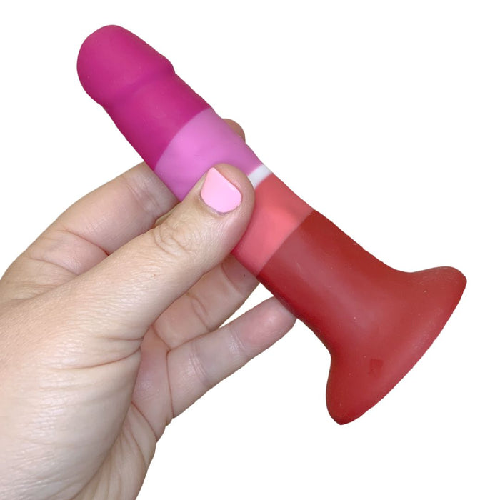 A light-skinned hand with pink nail polish holds the Lesbian Pride Small 5.5 Inch Silicone Suction Cup Dildo, featuring silky silicone in red, pink, and purple hues inspired by the Lesbian flag, against a white background.