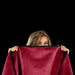 A person with long, light brown hair holds up a red Liberator Fascinator Throw King Size Waterproof Sex Blanket, covering their face up to their eyes, against a solid black background.
