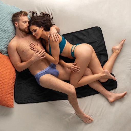 A man and woman in underwear cuddle closely on a bed atop the Liberator Fascinator Travel Sized Waterproof Sex Blanket, with colorful pillows nearby.