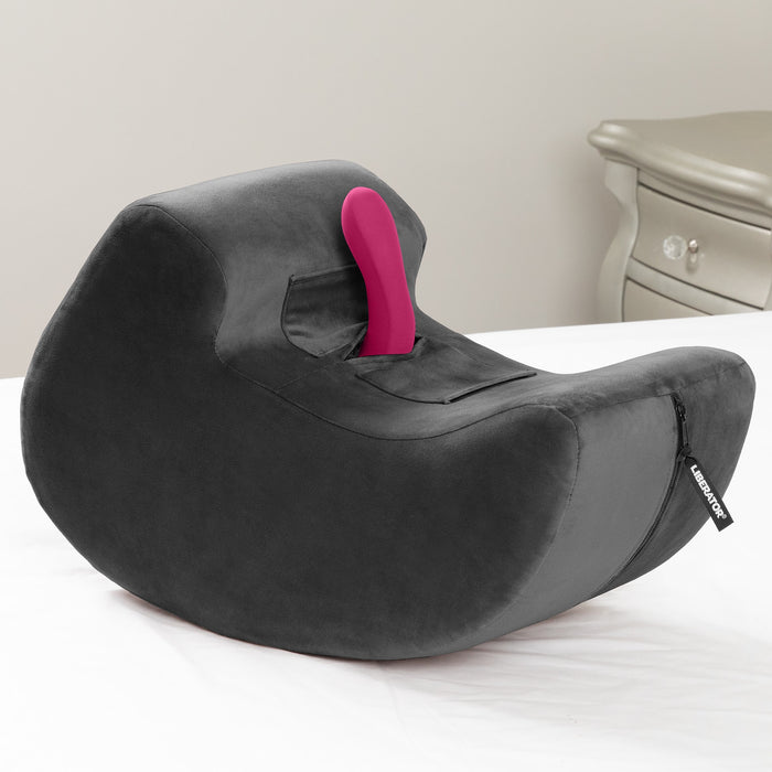The Liberator Pulse – Ergonomic Straddling Toy Mount in dark gray, featuring a pocket for your favorite toy, sits on a white surface next to a beige nightstand—designed for hands-free intimacy.