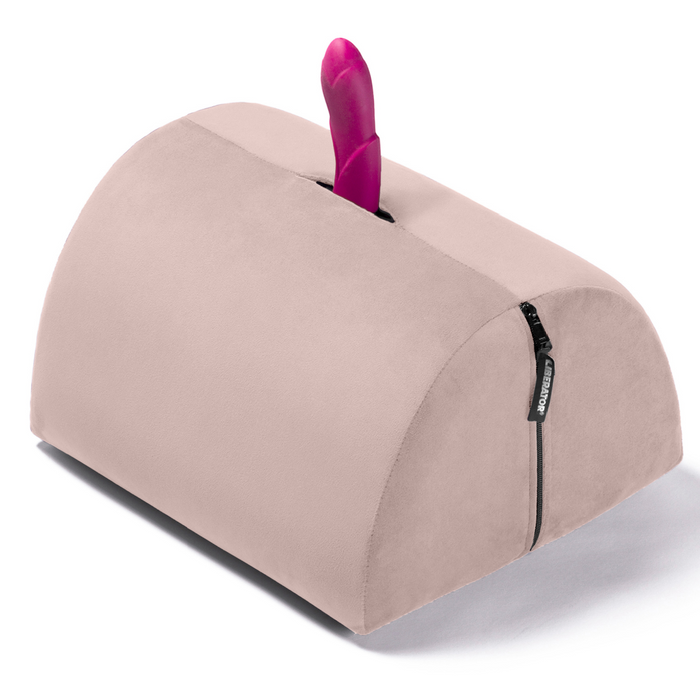 The Liberator BonBon Rideable Sex Toy Mount is a light pink, wedge-shaped foam pillow with a built-in slot for holding an upright vibrator and a black side zipper, offering discreet, hands-free pleasure.