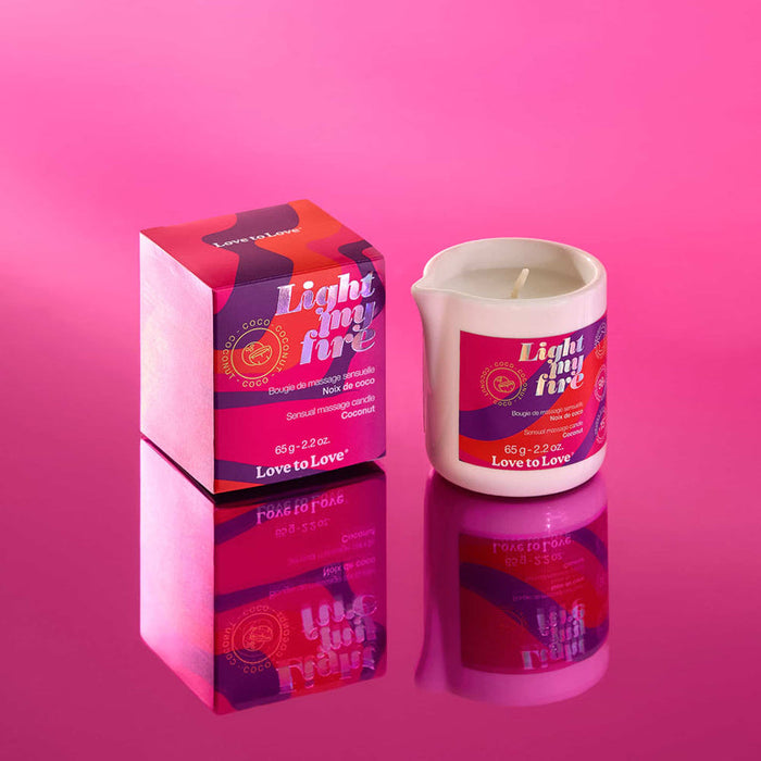 A white Light My Fire Coconut Scent Sensual Massage Candle sits in a small cup beside its matching pink and red box, both with bold graphics and the Love to Love brand, on a reflective pink surface.