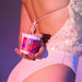 Dressed in sequined pale pink, someone holds the Light My Fire Lotus Flower Scent Sensual Massage Candle by their side, as purple and pink lights cast a soft, colorful glow.