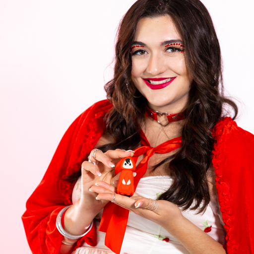 A smiling woman with wavy brown hair and bold red lipstick wears a red cape and white dress, holding the Little Red Discreet Powerful Bullet Vibrator, shaped like a small cloaked character, against a pinkish-white background.