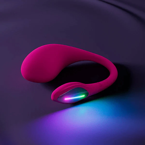 The Lovense Lush 4 Sound Activated Bluetooth Wearable Vibrator is pink silicone with a curved design for G-spot stimulation and a glowing multicolored light section, shown resting on a sleek dark surface.