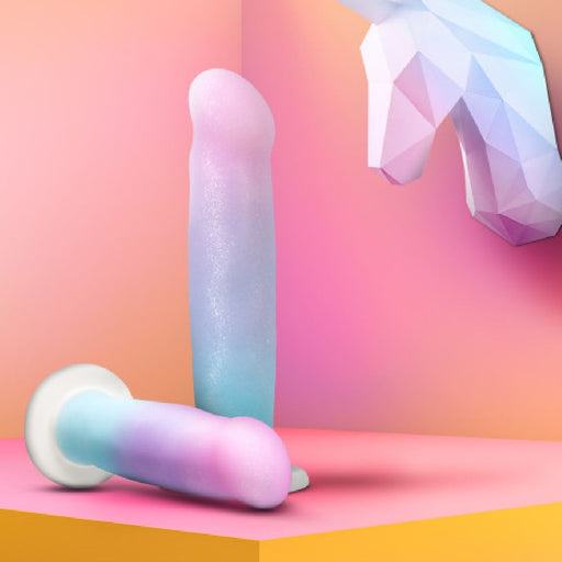 Two pastel-colored Lucky Sparkle Lightly Textured 8 Inch Silicone Suction Cup Dildos rest on a pink and orange surface, with a polygonal unicorn head in the corner and a gradient backdrop, emphasizing the magical vibe.