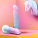 Two pastel-colored Lucky Sparkle Lightly Textured 8 Inch Silicone Suction Cup Dildos rest on a pink and orange surface, with a polygonal unicorn head in the corner and a gradient backdrop, emphasizing the magical vibe.
