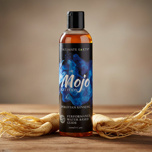 A bottle of Mojo Performance Water-Based Glide With Peruvian Ginseng sits on a wooden surface, flanked by two pieces of ginseng root.