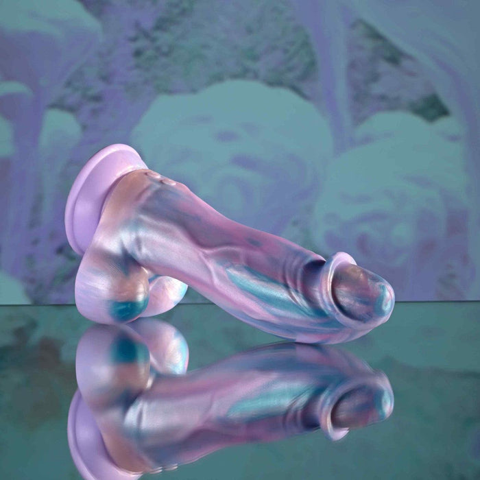 The Mythical Long Thick Vibrating Fantasy Dildo in Pastel Ombre, with pearlescent, iridescent hues and a sculpted design, rests on a reflective surface against a blue-purple abstract backdrop. Compatible with strap-on harnesses.
