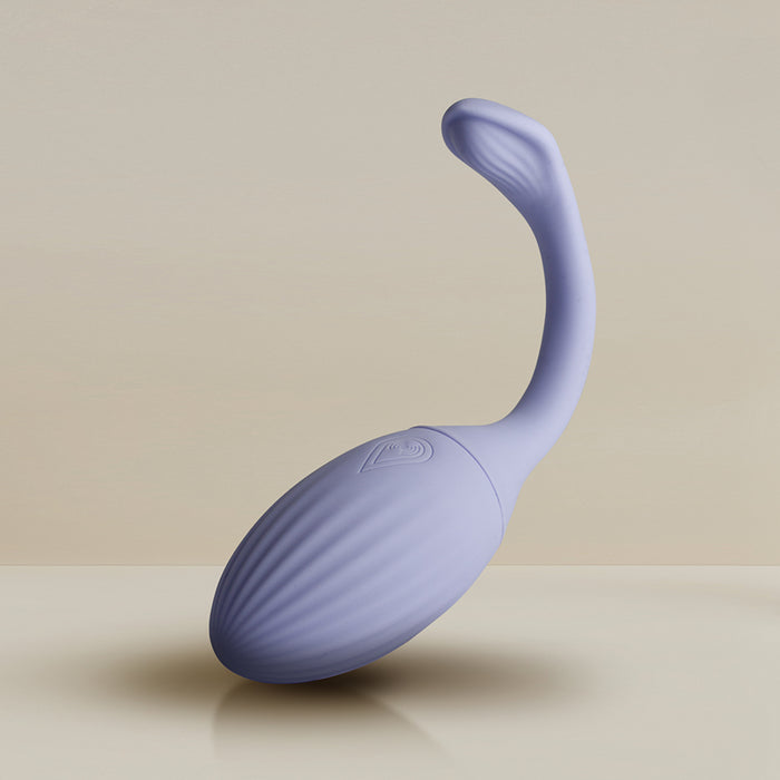 The Niya Rechargeable Kegel Exerciser and Vibrator with Remote, a purple silicone device featuring a ribbed oval base and long, curved handle for pelvic floor strengthening, is shown upright on a neutral surface against a beige background.