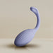 The Niya Rechargeable Kegel Exerciser and Vibrator with Remote, a purple silicone device featuring a ribbed oval base and long, curved handle for pelvic floor strengthening, is shown upright on a neutral surface against a beige background.