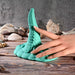 A realistic silicone hand touches the Octopus Tentacle Insertable Remote Controlled Grinder, a teal waterproof sex toy, on a wooden surface with assorted seashells and a gray stone background.