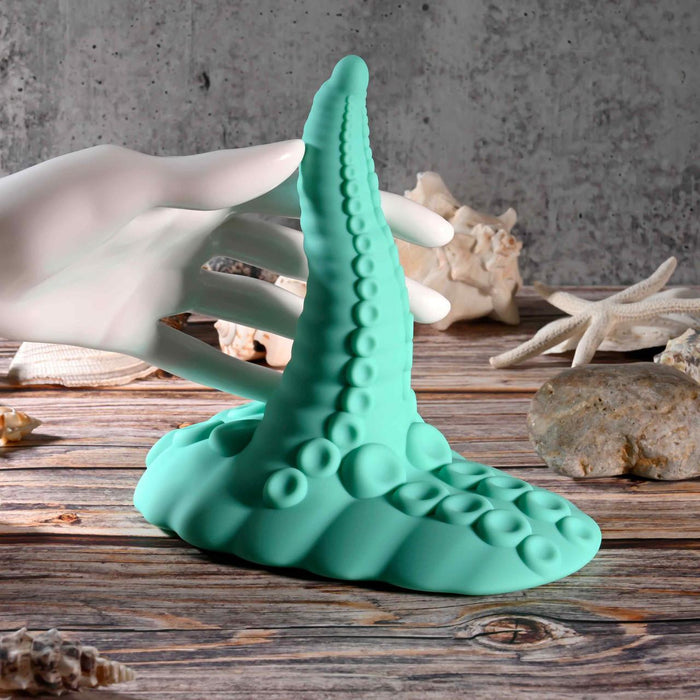 A white mannequin hand touches the teal Octopus Tentacle Insertable Remote Controlled Grinder on a rustic wooden surface, surrounded by seashells and starfish, with a gray textured wall in the background.
