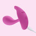 The Oly 2 Pressure Sensing App-Enabled Wearable Vibrator by Honey Play Box, in pink with a curved design, power button, and two indicator lights, is pictured charging via a white USB cable against a light pink background.