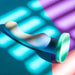 The Opal Dreams Slim 6 Inch Blue Ombre G Spot Dildo, a pastel silicone toy with a flared base for harness use, rests on a vibrant blue, teal, and purple background illuminated by soft, striped light patterns.