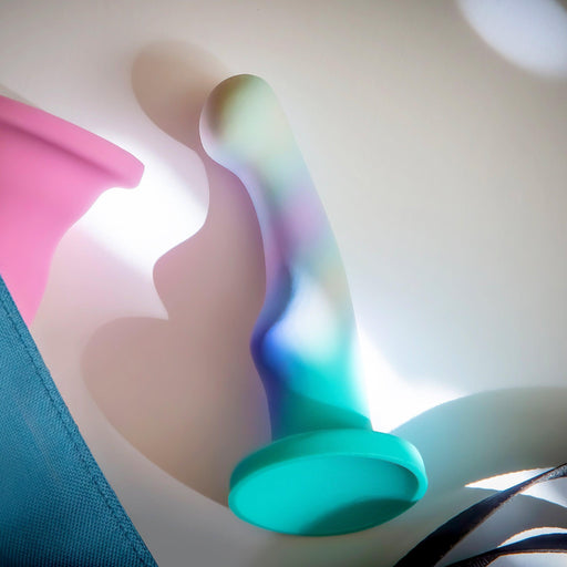 The Opal Dreams Slim 6 Inch Blue Ombre G Spot Dildo, a colorful silicone toy with a flared base for strap-on harness compatibility, lies on a white surface with light falling over it. Nearby are parts of a pink item and blue fabric.