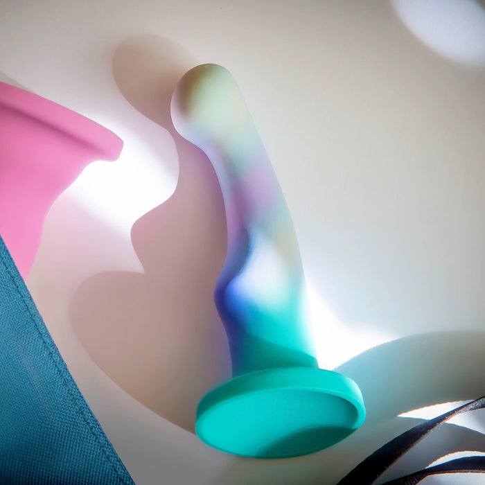 The Opal Dreams Slim 6 Inch Blue Ombre G Spot Dildo, a colorful silicone toy with a flared base for strap-on harness compatibility, lies on a white surface with light falling over it. Nearby are parts of a pink item and blue fabric.