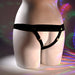 A mannequin torso showcases the Our First Hollow Strap-On Dildo + Harness Set - Vanilla, a minimal black harness-style lingerie piece compatible with a hollow strap-on, against a backdrop with colorful light reflections.