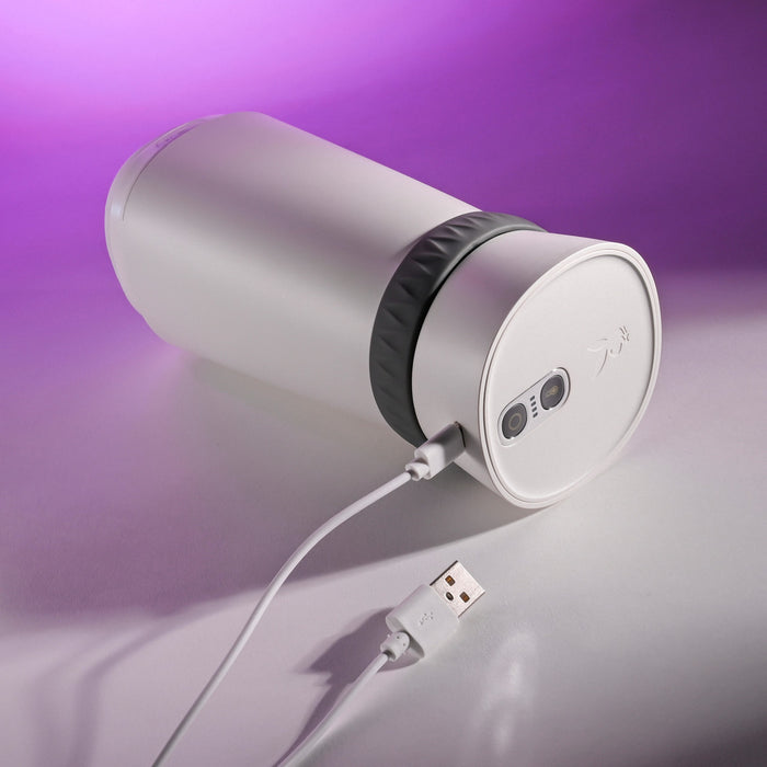The Playboy Twist & Stroke Heated Vibrating Stroker, a cylindrical white device with a gray band, lies on its side on a pink and purple surface with a USB cable plugged into its base.