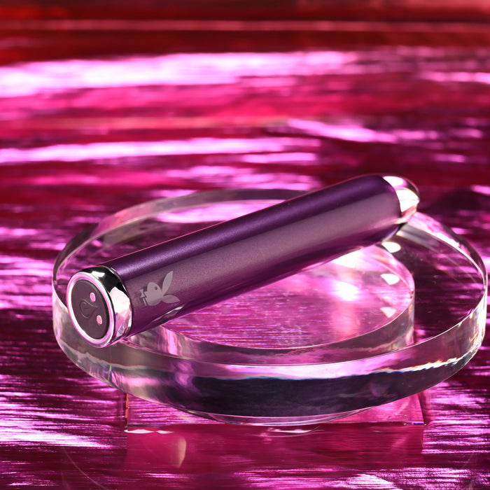 The Amethyst Slim Waterproof Rechargeable Glass Vibrator, with chrome-colored ends and an engraved bunny logo near its base, rests on a clear circular platform against a shiny pink background.